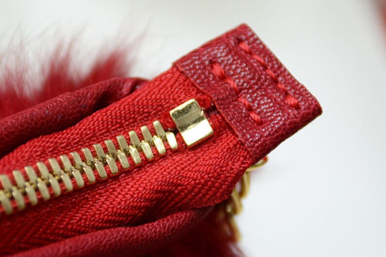 May include: Close-up of a red fabric with a gold zipper. The zipper is partially open, revealing the gold teeth and a gold zipper pull. The fabric has a textured appearance and is stitched with red thread. A small amount of red fur is visible.