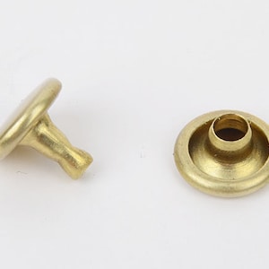 May include: Two brass rivets, one with a rounded head and post, the other a hollow base. The rivets are a shiny gold color and are likely used for fastening materials together. They are commonly used in leathercraft and garment construction.