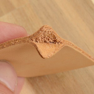 May include: Close-up of a piece of light brown leather, folded to create a pointed shape. The edges of the leather are visible, showing the layered construction. The leather has a textured, natural appearance.