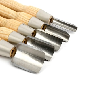 May include: A collection of wood carving tools featuring stainless steel blades. The tools have light brown wooden handles and are arranged diagonally against a white backdrop. The blades have a curved, scoop-like form.