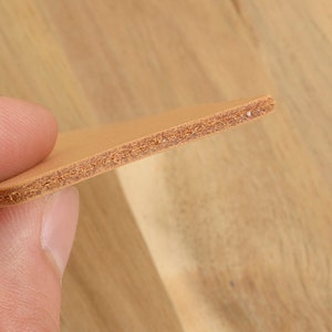 May include: Close-up of a piece of tan leather. The leather is held between fingers, showcasing its layered texture and light brown color. The edges reveal a detailed cross-section of the material, highlighting its construction.