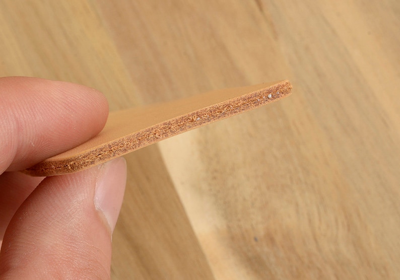 May include: Close-up of a piece of tan leather. The leather is held between fingers, showcasing its layered texture and light brown color. The edges reveal a detailed cross-section of the material, highlighting its construction.