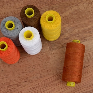 Puede incluir: Una colección de coloridas bobinas de hilo en varios tonos, incluyendo naranja, blanco, amarillo, gris y marrón. Cada bobina tiene un centro amarillo y está dispuesta sobre una superficie de madera. El hilo es probablemente para coser o manualidades.