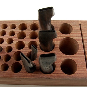 Puede incluir: Un organizador de herramientas de madera con agujeros y ranuras de varios tamaños. Varias herramientas de metal están insertadas en los agujeros. La madera es de color marrón rojizo.