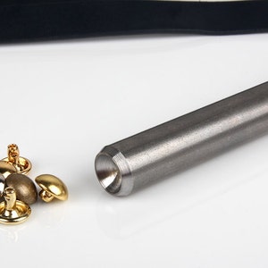 May include: A collection of metal rivets in various finishes, including gold, silver, and bronze, alongside a cylindrical metal tool. The rivets have rounded heads and are designed for fastening. The tool is a long, gray metal rod.