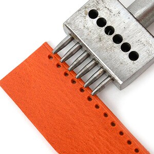 May include: A metal leather punch tool creating holes in a piece of orange leather. The tool has multiple pointed punches. The leather is a bright orange colour, and the holes are evenly spaced. The leather is approximately 10 cm wide.