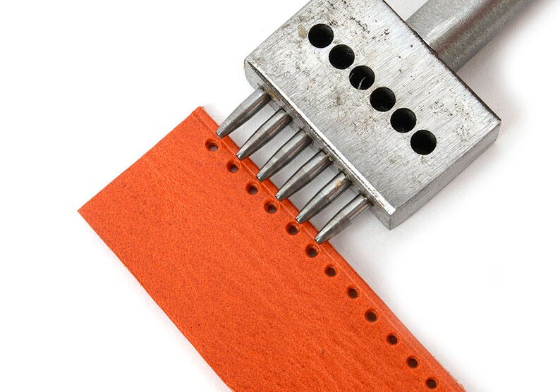 May include: A metal leather punch tool creating holes in a piece of orange leather. The tool has multiple pointed punches. The leather is a bright orange colour, and the holes are evenly spaced. The leather is approximately 10 cm wide.