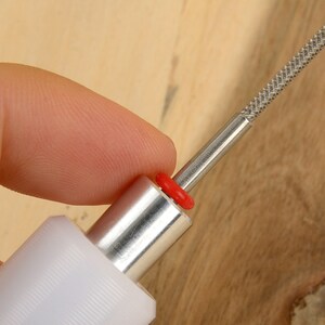 May include: A close-up of a metal and plastic tool with a textured, silver-colored tip. The tool has a white handle, a silver shaft, and a red ring. The tool is held by a person's finger.
