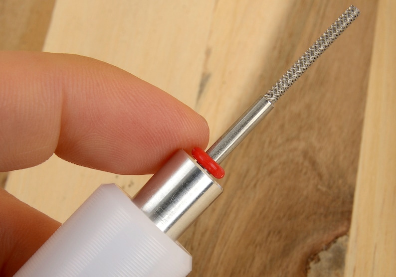 May include: A close-up of a metal and plastic tool with a textured, silver-colored tip. The tool has a white handle, a silver shaft, and a red ring. The tool is held by a person's finger.