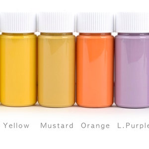 May include: Six small bottles of paint in a row, each filled with a different color. The colors are yellow, mustard, orange, light purple, and purple. Each bottle has a white cap, and the names of the colors are printed below.
