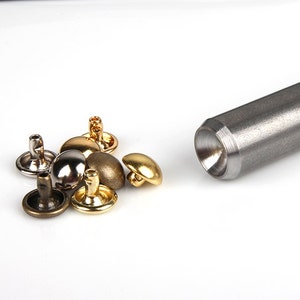May include: A collection of metal rivets in various finishes, including gold, silver, and bronze, alongside a silver metal setting tool. The rivets have a rounded, domed top and a flat base. The tool is cylindrical with a concave end.