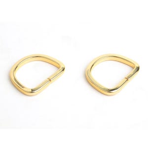 May include: Two gold-colored D-rings, each with a flat, slightly curved design. The rings are made of a shiny metal and are positioned against a white background. These rings are likely used for bag making or other craft projects.