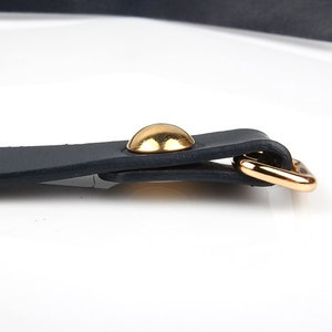 May include: Close-up of a black leather strap with a gold-colored metal buckle and a gold-colored rivet. The strap is likely part of a belt or accessory. The background is white.