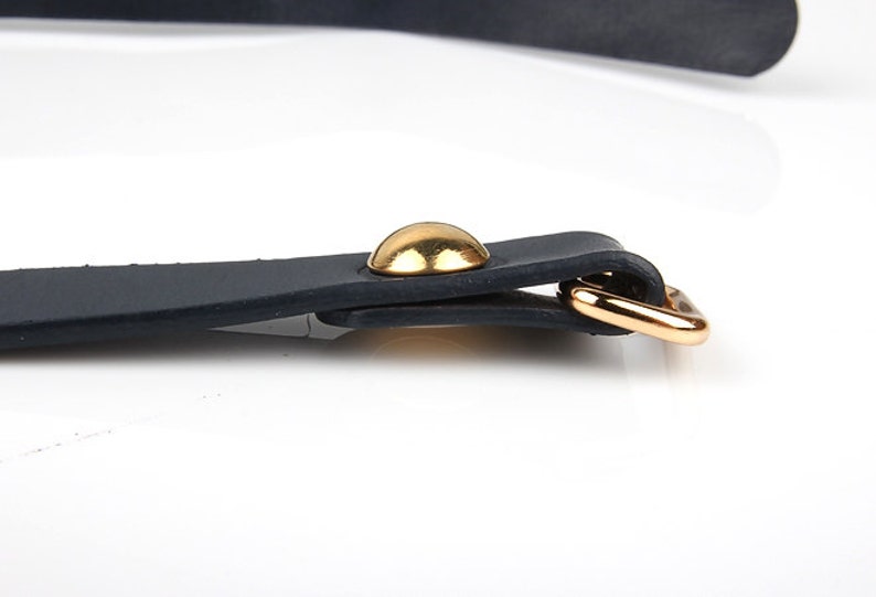May include: Close-up of a black leather strap with a gold-colored metal buckle and a gold-colored rivet. The strap is likely part of a belt or accessory. The background is white.