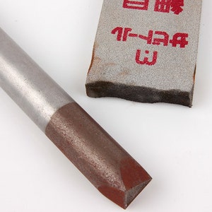 May include: Close-up of a metal chisel with a brown cutting edge and a gray handle. A gray sanding block with Japanese text is in the background. The tools are on a white background.