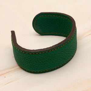 May include: A green leather cuff bracelet with brown edges and gold stitching. The bracelet has a textured surface and a curved design. This accessory is suitable for everyday wear. The bracelet is approximately 2 cm wide.