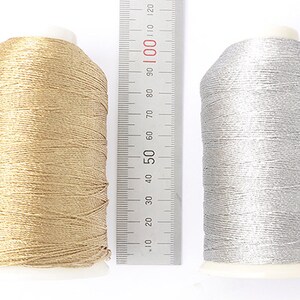 May include: Two spools of metallic thread, one gold and one silver, sit side-by-side. A ruler with inch and centimeter markings is placed between them. The gold spool is approximately 4 inches tall, and the silver spool is slightly shorter.