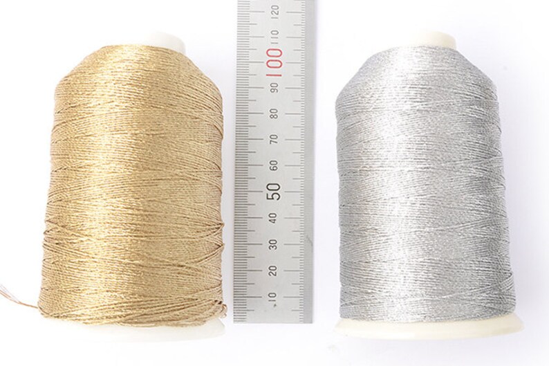 May include: Two spools of metallic thread, one gold and one silver, sit side-by-side. A ruler with inch and centimeter markings is placed between them. The gold spool is approximately 4 inches tall, and the silver spool is slightly shorter.