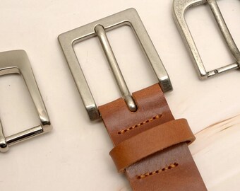 May include: Close-up of three silver-colored belt buckles. The central buckle is attached to a brown leather belt. The buckles have a rectangular and rounded design. The leather belt is stitched with yellow thread.