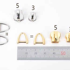 May include: A collection of metal hardware components, including silver and gold-toned zipper pulls and D-rings. The image includes a ruler for scale, with the zipper pulls labeled as sizes 3 and 5.