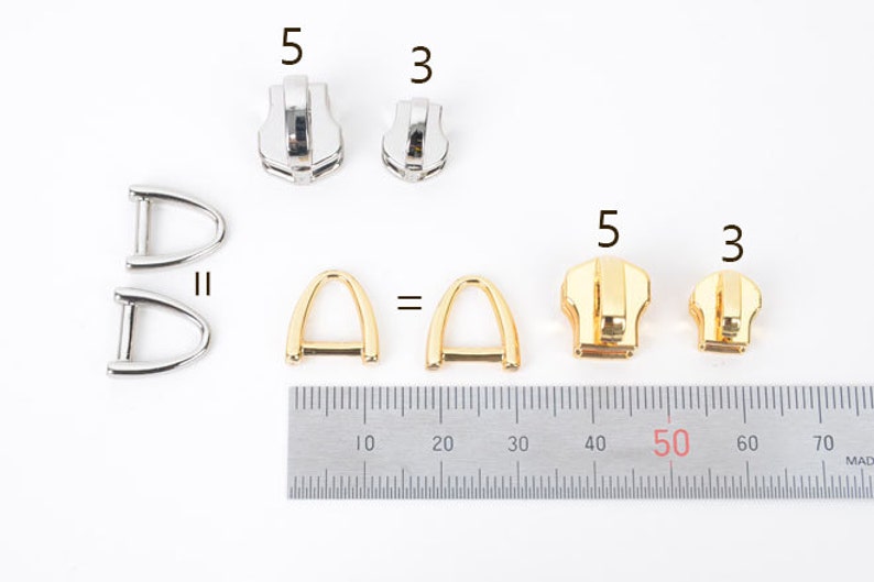 May include: A collection of metal hardware components, including silver and gold-toned zipper pulls and D-rings. The image includes a ruler for scale, with the zipper pulls labeled as sizes 3 and 5.