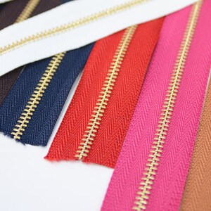 Puede incluir: Primer plano de varias cremalleras coloridas con dientes dorados. Las cremalleras son de varios colores, incluyendo blanco, azul marino, rojo, rosa y marr&oacute;n. Est&aacute;n dispuestas en diagonal, mostrando su dise&ntilde;o y variaciones de color.