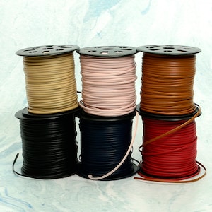 May include: A collection of spools of leather cord in various colors, including tan, pink, brown, black, navy, and red. The cords are neatly wound on black plastic spools, showcasing the different hues and textures of the material. The cords are likely used for jewelry making or crafting.
