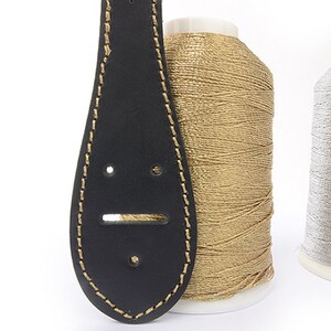 May include: A black leather guitar strap with gold stitching and a gold thread spool next to a silver thread spool. The strap has a unique teardrop shape with two small holes and a larger rectangular hole.