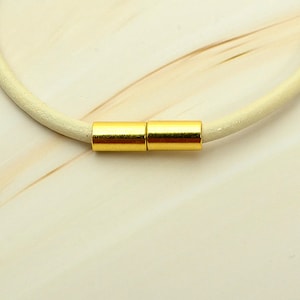 May include: A cream-colored leather necklace with a gold-colored magnetic clasp. The clasp is made of two cylindrical pieces that connect to form a secure closure. The necklace is simple and elegant.