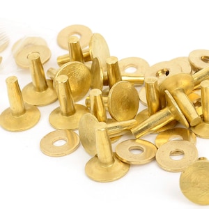 Puede incluir: Un conjunto de remaches y arandelas metálicas de color dorado brillante. Los remaches tienen un vástago cilíndrico y una cabeza plana y redonda. Las arandelas también son redondas con un agujero en el centro. Estas piezas son probablemente para marroquinería u otros proyectos de bricolaje.