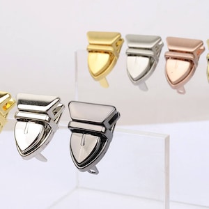 Pode incluir: Uma coleção de fechos de metal para bolsas em vários acabamentos, incluindo dourado, prateado e ouro rosa. Os fechos têm formato de escudo com uma parte superior articulada e um pequeno laço na parte inferior. Exibidos em uma superfície acrílica transparente.