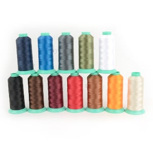 Könnte beinhalten: Eine Sammlung von zwölf Garnrollen in verschiedenen Farben, darunter Marineblau, Blau, Grau, Olivgrün, Weiß, Schwarz, Braun, Burgunderrot, Rot, Orange und Creme. Jede Rolle ist auf einer hellgrünen Basis montiert, geeignet für Näh- und Bastelprojekte.