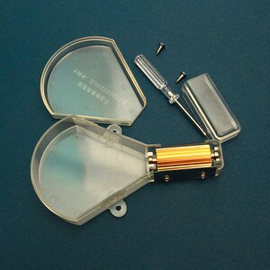 Puede incluir: Un dispositivo de pl&aacute;stico transparente desmontado con un componente dorado, un peque&ntilde;o destornillador y dos tornillos. El dispositivo tiene forma semicircular con una secci&oacute;n rectangular. El fondo es de color verde azulado oscuro.