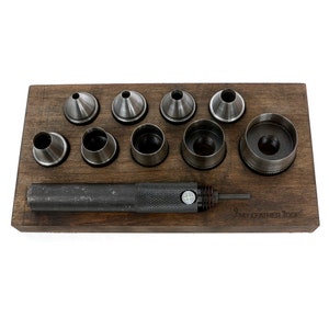 May include: A set of metal hole punch tools arranged on a wooden base. The set includes various sizes of punch heads and a handle. The wooden base has the text "MY LEATHER TOOL" etched on it. The tools are designed for leathercraft.