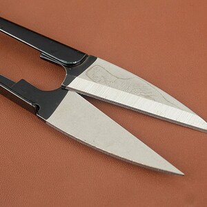K&ouml;nnte beinhalten: Nahaufnahme einer Schere mit schwarzen Griffen und silbernen Klingen. Die Klingen sind scharf und abgewinkelt, mit einem dekorativen Muster in der N&auml;he des Drehpunktes. Die Schere liegt auf einem braunen Hintergrund.