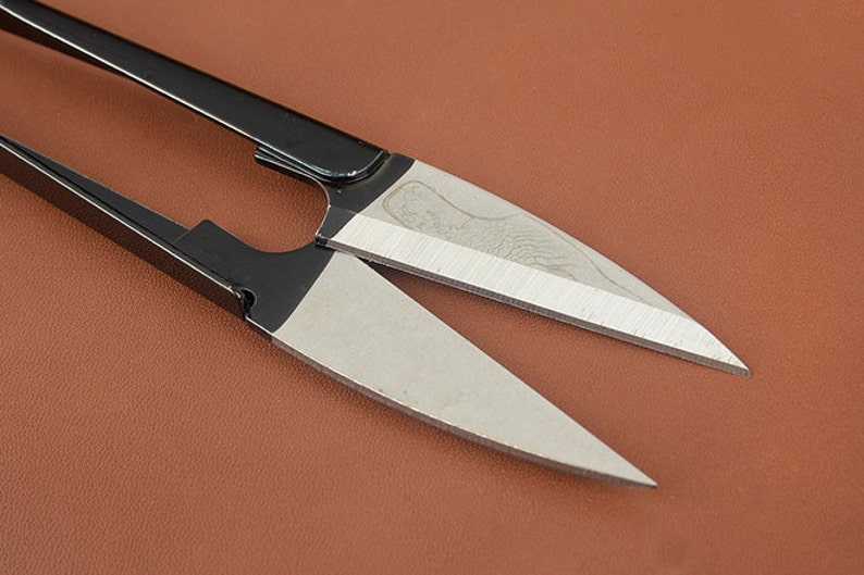 K&ouml;nnte beinhalten: Nahaufnahme einer Schere mit schwarzen Griffen und silbernen Klingen. Die Klingen sind scharf und abgewinkelt, mit einem dekorativen Muster in der N&auml;he des Drehpunktes. Die Schere liegt auf einem braunen Hintergrund.