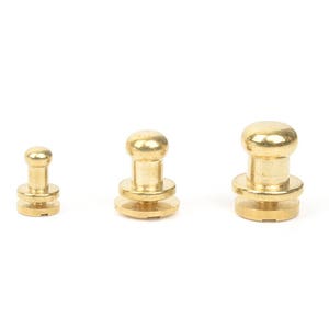 Puede incluir: Tres remaches de latón de diferentes tamaños sobre un fondo blanco. Cada remache tiene una cabeza redondeada y una base con un diseño circular. Estos sujetadores se utilizan comúnmente en marroquinería y otros proyectos de artesanía.