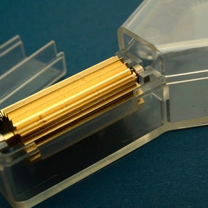 Puede incluir: Primer plano de un objeto cil&iacute;ndrico dorado y estriado, parcialmente encerrado en un recipiente de pl&aacute;stico transparente. El objeto tiene una serie de estr&iacute;as paralelas a lo largo de su longitud. El recipiente es rectangular con tapa abatible. El fondo es de color verde azulado.