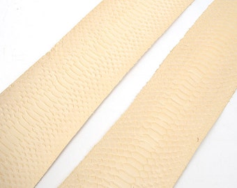 May include: Two strips of light beige leather with a reptile scale pattern. The leather is textured and appears to be a natural material. The strips are angled across a white background.
