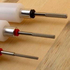 May include: Close-up of three white cylindrical tools with metal shafts and textured handles. The tools have a silver metal collar and a black or red rubber ring. The background is a light brown wooden surface.