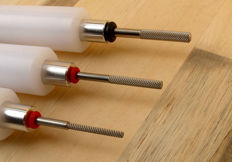 May include: Close-up of three white cylindrical tools with metal shafts and textured handles. The tools have a silver metal collar and a black or red rubber ring. The background is a light brown wooden surface.