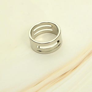 Può includere: Un anello color argento dal design moderno. L'anello presenta tre sezioni orizzontali ritagliate attorno alla fascia. L'anello è realizzato in un materiale metallico lucido.