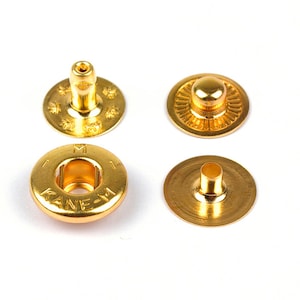 May include: Four gold-coloured metal snap fasteners. Two are complete snaps, and two are individual pieces. One piece has the text "KANE-M" around the centre hole. The fasteners are on a white background.