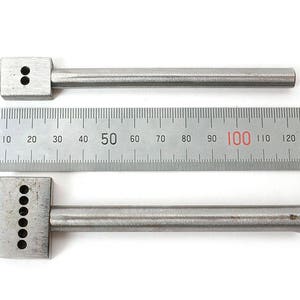 May include: Two silver leather working tools with multiple prongs and a ruler. The tools have a rectangular head with prongs and a cylindrical handle. The ruler is marked in centimetres.