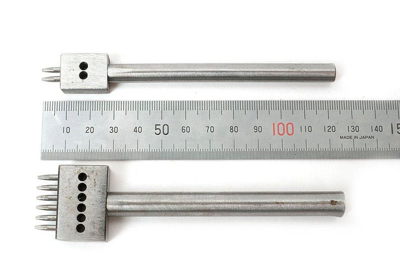 May include: Two silver leather working tools with multiple prongs and a ruler. The tools have a rectangular head with prongs and a cylindrical handle. The ruler is marked in centimetres.