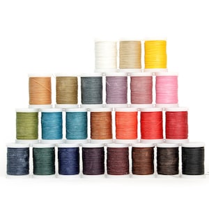 Puede incluir: Una colección de coloridas bobinas de hilo, dispuestas en forma de pirámide. Los colores del hilo incluyen blanco, beige, amarillo, naranja, rosa, verde, azul, marrón y negro. Cada bobina está sobre una base de plástico blanco, ideal para manualidades.