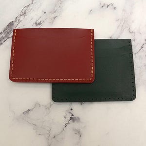 Puede incluir: Dos tarjeteros de cuero. Uno es de color rojo intenso con costuras color crema, y el otro es verde oscuro con costuras a juego. Ambos son rectangulares con un bolsillo en un lado.