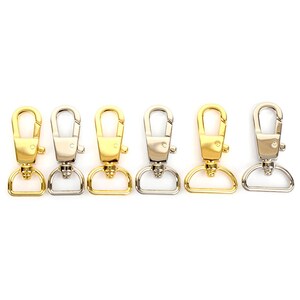 May include: A row of five metal swivel clasps in gold and silver tones. Each clasp has a D-ring base and a spring-loaded hook. The clasps are arranged horizontally against a white background.