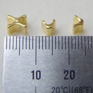 May include: Three small, gold-colored metal beads in different shapes are displayed next to a ruler. The beads are a cross, a U-shape, and a curved shape. The ruler shows measurements in inches and the temperature is 20&deg;C (68&deg;F).
