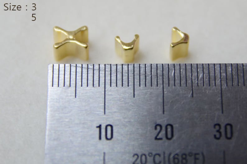 May include: Three small, gold-colored metal beads in different shapes are displayed next to a ruler. The beads are a cross, a U-shape, and a curved shape. The ruler shows measurements in inches and the temperature is 20&deg;C (68&deg;F).